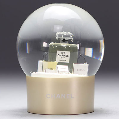 Boule à neige CHANEL en verre représentant une bouteille du parfum CHANEL n°5 entourée de sacs cadeaux ornés du logo CHANEL