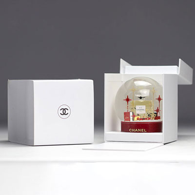 Boule à neige CHANEL en verre représentant une grande bouteille du parfum CHANEL n°5, entourée de paquets cadeaux ornés du logo CHANEL en rouge et d'étoiles